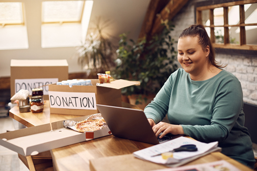 Happy non profit staff sending volunteer recruitment email using laptop with donation boxes beside her